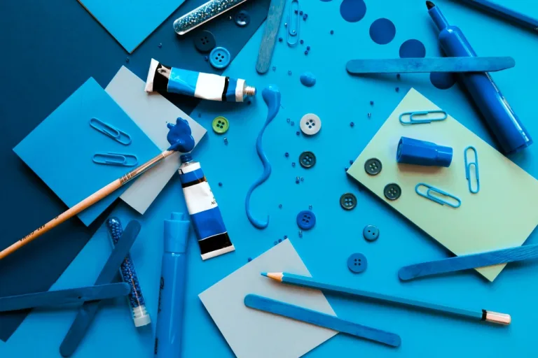 Blue craft supplies including paint tubes, buttons, paper clips and colored paper laid flat
