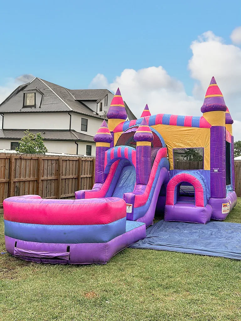 Pink and purple castle bounce house with slide set up in a fenced backyard