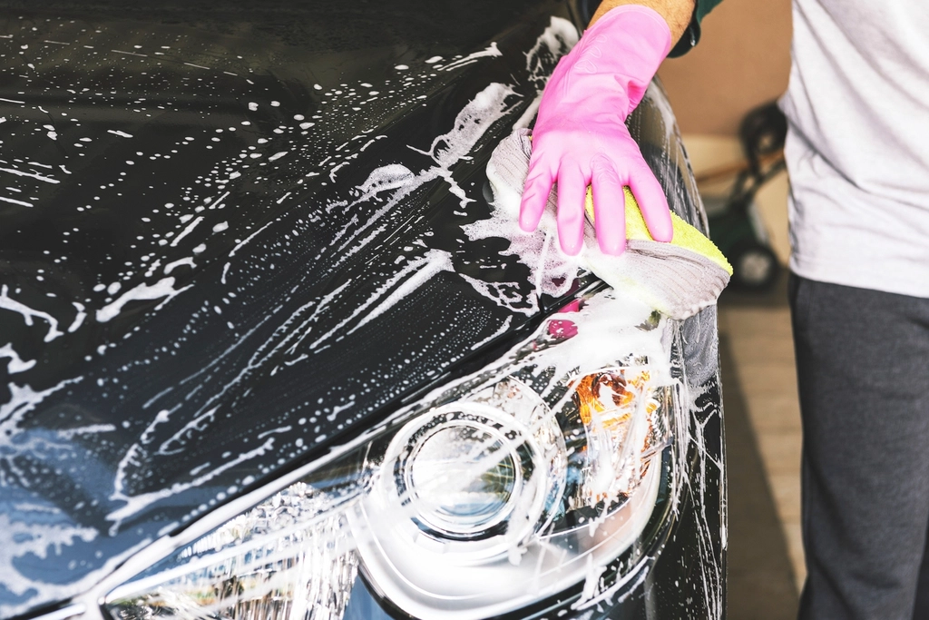 Gloved hand washing a dark car near the headlight with a soapy sponge