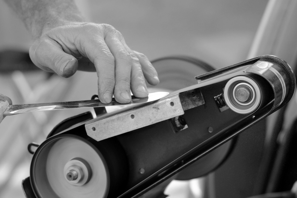 Black-and-white close-up of a hand sharpening a knife on a belt grinder