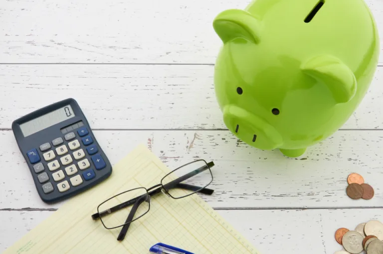 Green piggy bank next to a calculator, eyeglasses and coins on a white table