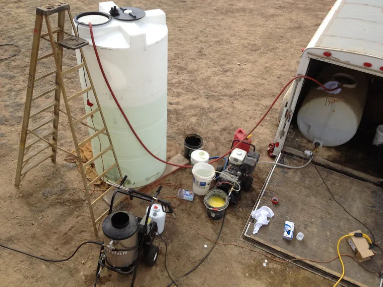 Pressure washing setup with a water tank, hoses, a gas-powered washer and a ladder next to a work trailer