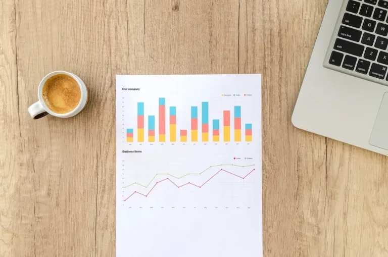 Printed sales charts next to a laptop and coffee on a wooden desk