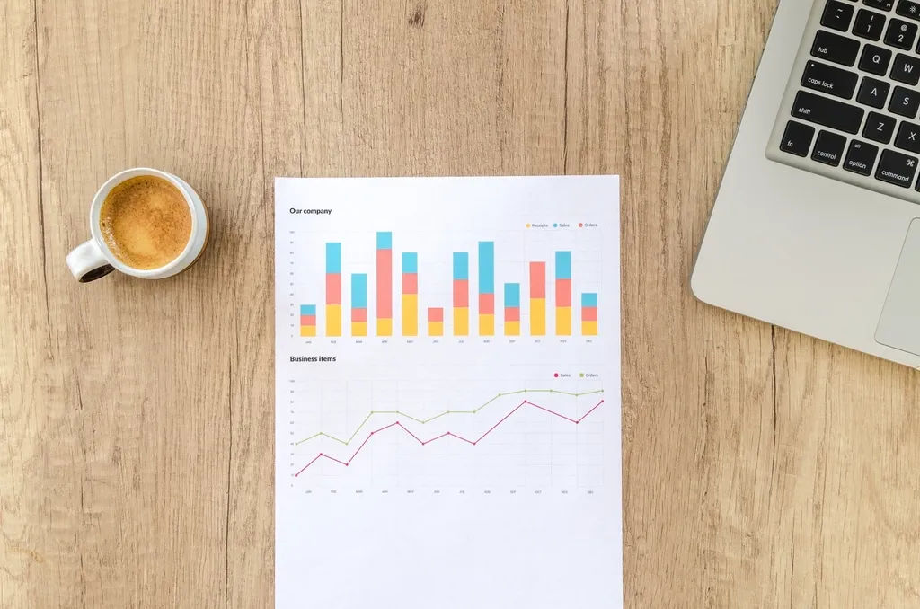 Printed sales charts next to a laptop and coffee on a wooden desk