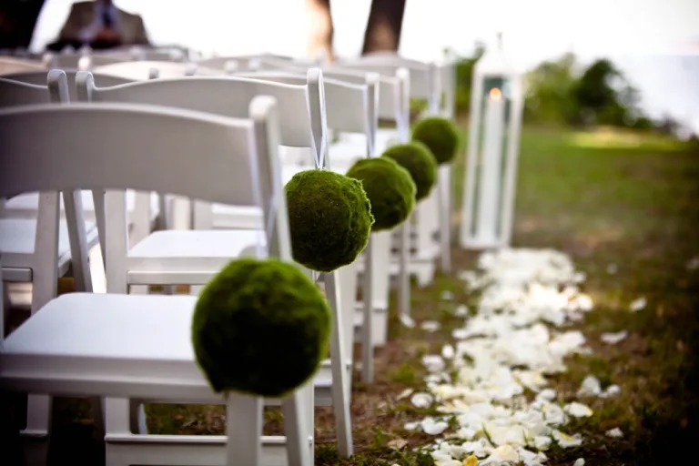 White ceremony chairs lining an outdoor aisle decorated with moss balls and petals