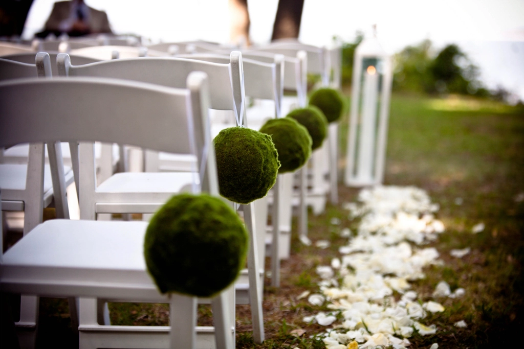 White ceremony chairs lining an outdoor aisle decorated with moss balls and petals
