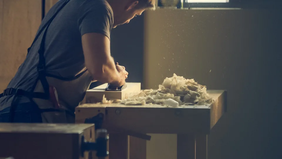 Woodworker planing a piece of wood at a workbench with shavings piled up