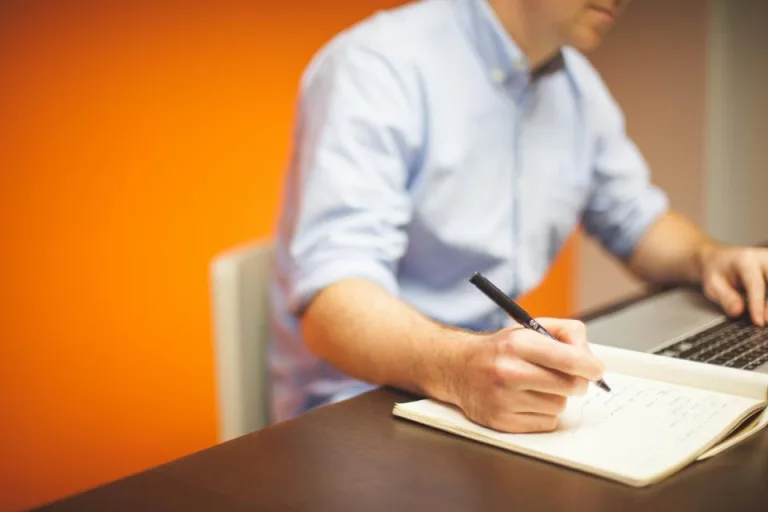 Person writing notes in a notebook beside a laptop at a desk